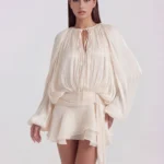 Front mid-length view of The Giovannina Luminous Pleated Ruffled Mini Dress in cream, highlighting the keyhole neck tie, long blouson sleeves, and draped twist-front sash waist.