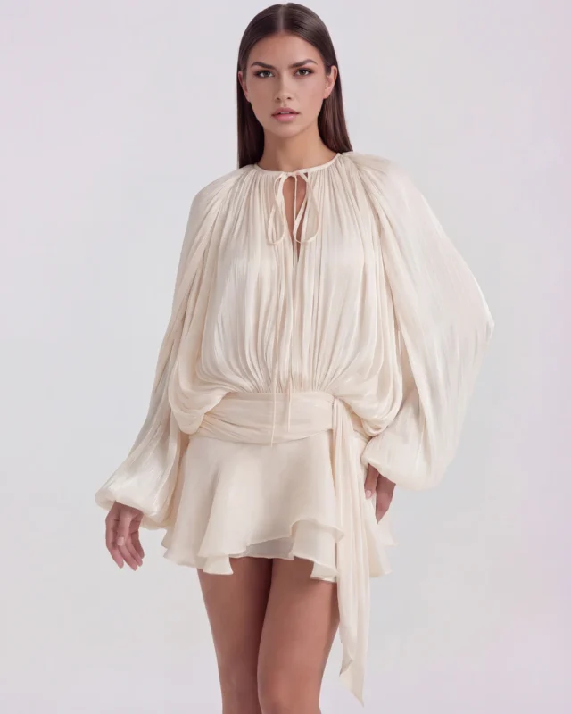 Front mid-length view of The Giovannina Luminous Pleated Ruffled Mini Dress in cream, highlighting the keyhole neck tie, long blouson sleeves, and draped twist-front sash waist.