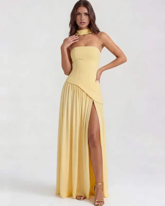 Front portrait of the light yellow corset evening dress, displaying the sharp diagonal waist seam, a dramatic high thigh split, and matching gold high-heeled strappy sandals.