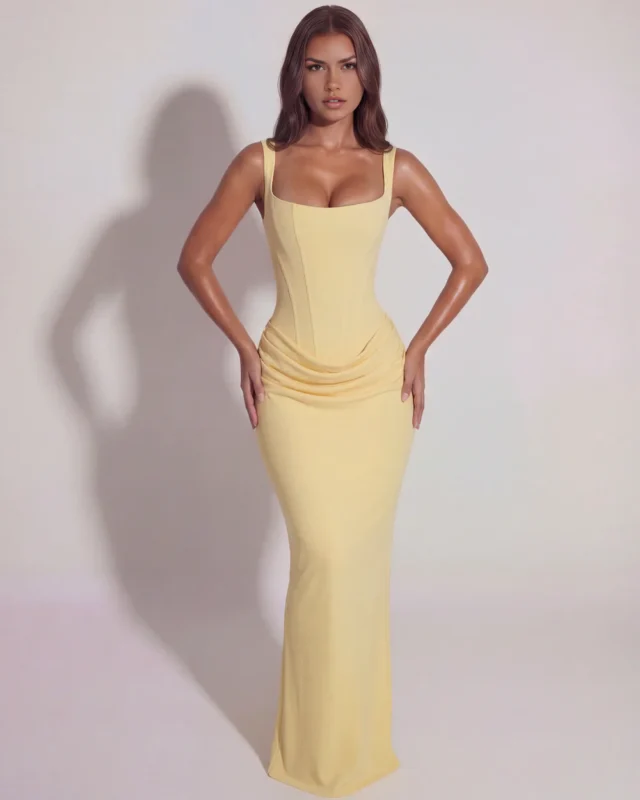 Front full-length view of the Eleonor structured corset maxi dress in a soft pastel butter yellow, showcasing the clean square neckline and elegant cowl draping along the drop-waist hips with a hands-on-hips pose.