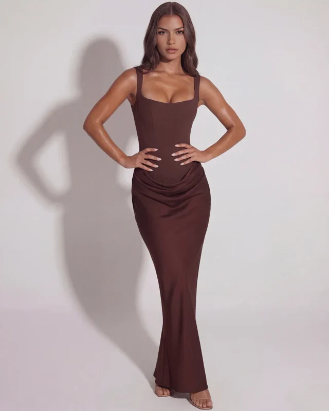 Full-length front perspective of a deep chocolate brown satin corset dress with a hands-on-hips pose, emphasizing the dramatic hourglass contouring and premium event wear styling.