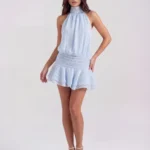 Front full-length studio portrait of the Ischia high-neck halter mini dress in light ice blue, showcasing a blouson-style pleated bodice, a wide smocked drop-waist band, and a flirty tiered ruffle skirt.