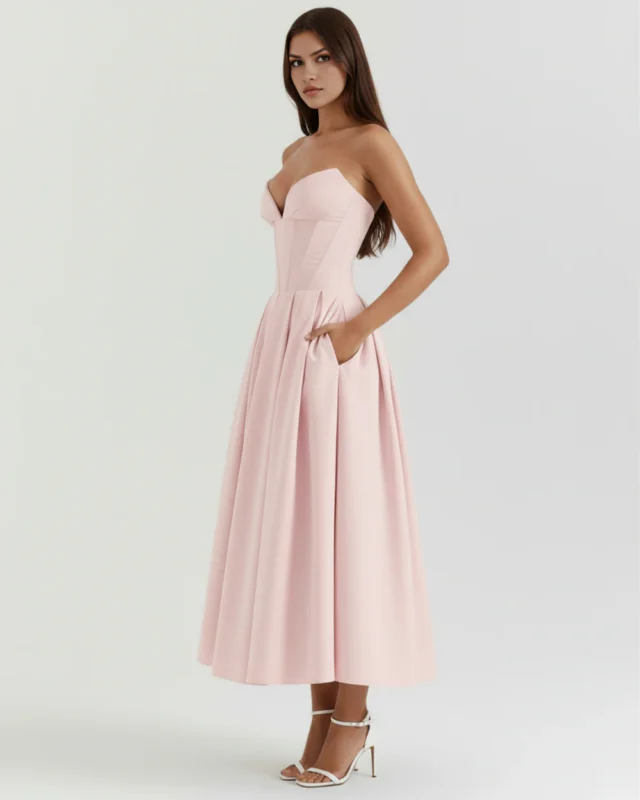 Three-quarter full-body profile view of the model posing with one hand in her pocket wearing the blush pink strapless evening midi dress, highlighting the tea-length hemline and defined waist panels.
