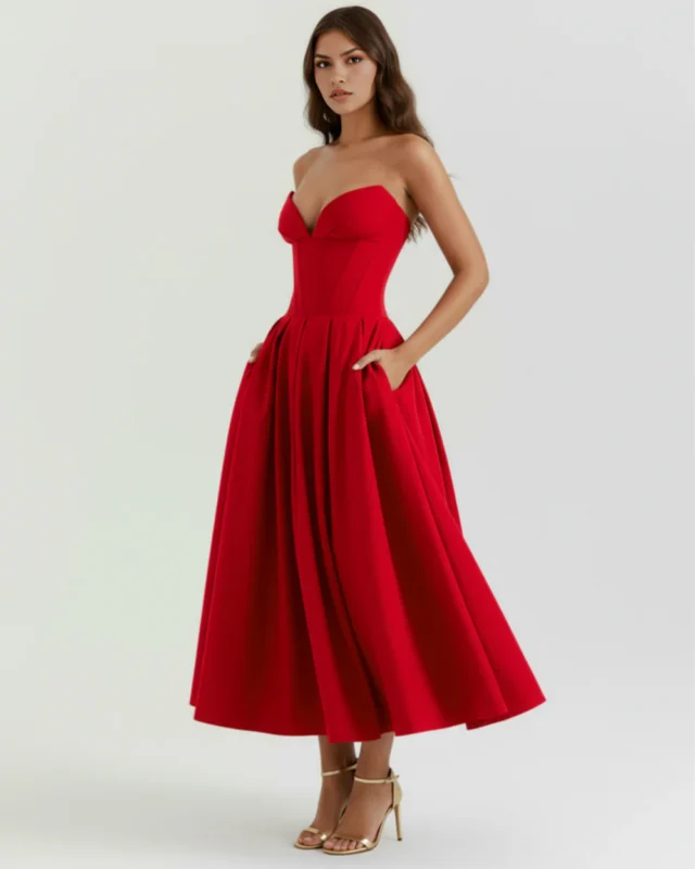 Full-body side profile shot of the model wearing the bright red strapless corset midi dress with hands in her pockets, highlighting the architectural volume of the flared tea-length skirt.