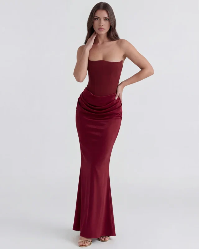 Front full-length shot of the Rosalinda strapless corset maxi gown in deep burgundy red, highlighting the hand-on-neck pose, sculpted pointed neckline, and fluid satin drop-waist draping.