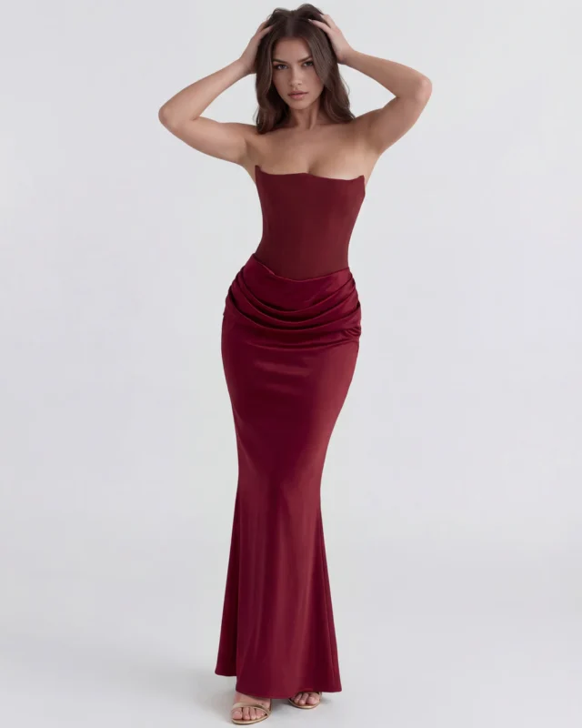 Full-length front perspective of a rich wine burgundy strapless gown with hands-on-head styling, showcasing the body-contouring bodice panels and dramatic cascading maxi skirt.