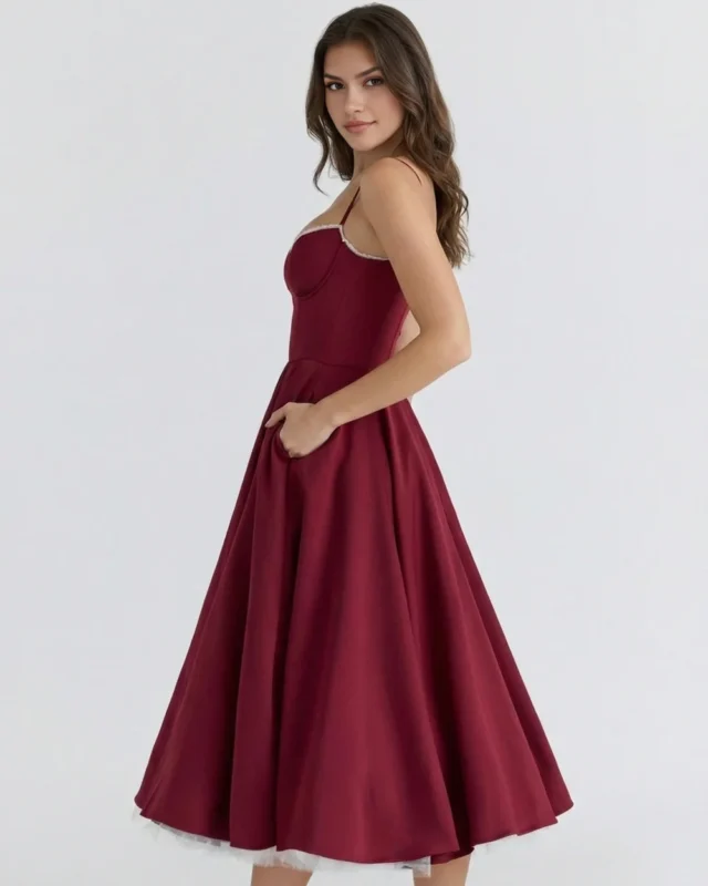 Side profile full-length view of a deep burgundy red A-line formal dress with hands in pockets, showcasing the smooth curve-defining fit and premium heavy satin-feel drapes.