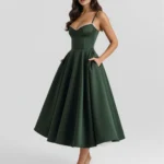 Three-quarter front full-length perspective of the Selene midi dress in deep forest green, emphasizing the hands-in-pockets pose, a supportive sweetheart bustier bodice, and white stiletto heels.