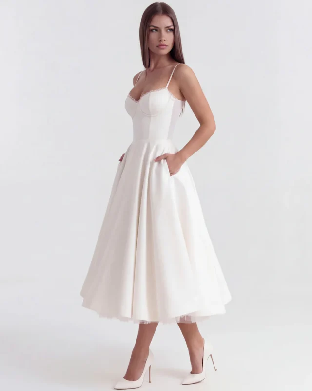 Side profile full-length perspective of the white corseted midi bridal gown with hands in functional side pockets, highlighting the structured waistline, white stiletto heels, and elegant satin finish.